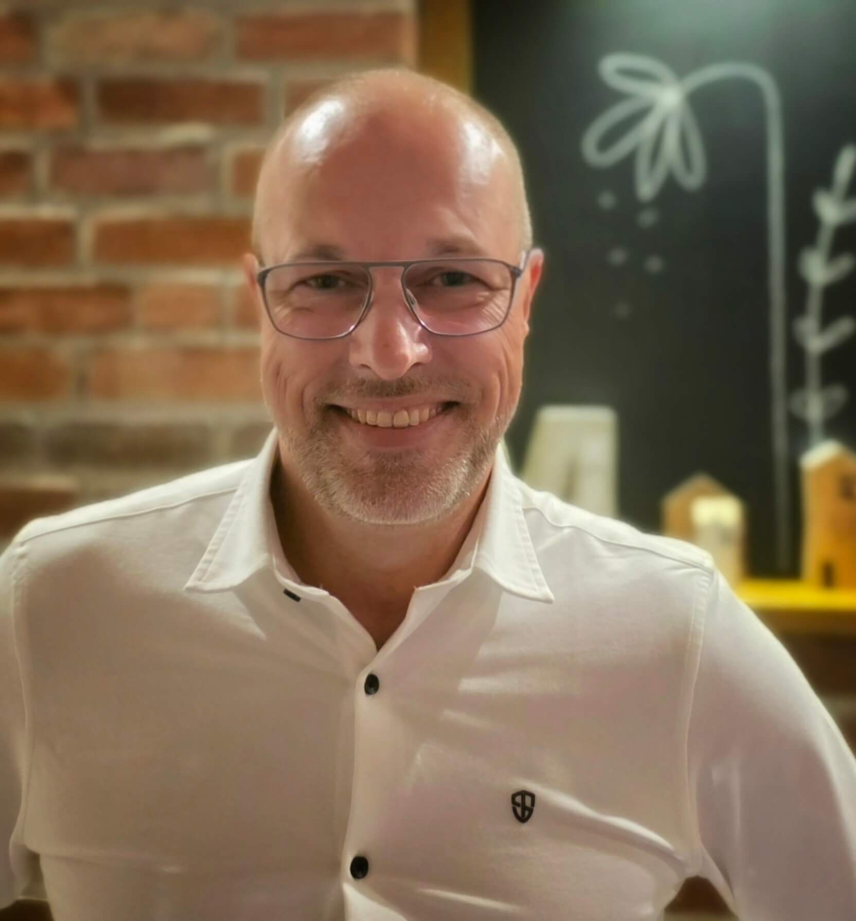 Mann mit Brille und Bart, der ein weißes Hemd trägt und lächelt. Im Hintergrund sind eine gemusterte Wand und eine Tafel mit Zeichnungen sichtbar.